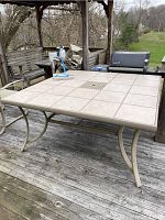 Square patio table with beige tile surface on a wooden deck, showing the full table and metal legs.