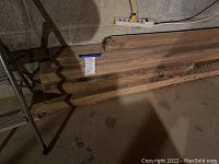 Stack of eight large oak wood planks leaning against a concrete block wall, showing length and texture of wood.