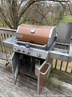 Front view of grill on deck showing lid closed, control knobs, and cabinet doors open revealing propane tank