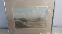 Framed watercolour painting showing landscape scene with hills and sky, artist signature visible at bottom right.