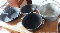 Overview of cast iron pans and pots along with aluminum pot displayed on a wooden surface, showing size and condition.