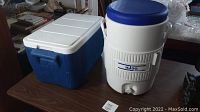 Full view of blue and white rectangular Coleman cooler and white cylindrical 5 gallon Igloo drink dispenser with blue lid and spigot on a table.