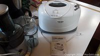 Black & Decker bread maker with open lid showing bread pan and instruction manual beside it, placed on a white surface.