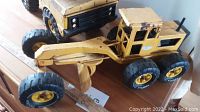 Angle view of yellow steel grader toy showing details of blade, tires, and cab with wear and paint chipping.
