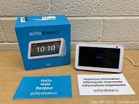 Photo showing Amazon Echo Show 5 device turned on with echo logo visible, next to the product packaging box and printed user guides.