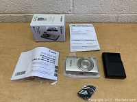Photo of Canon PowerShot ELPH 180 camera, manual, charger, USB cable sealed in plastic, box and documentation on table against beige wall