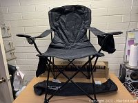 Front view of black Coleman folding chair with padded backrest, armrests, cup holder, small pouch, and carrying bag placed underneath on table.