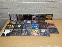 Top-down view of 22 music CDs laid out in jewel cases on a table showing album covers.