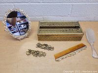 Photo shows a front view of vintage bathroom items including ornate oval mirror with gold-tone frame, metal filigree designed jewelry box, tortoiseshell comb with metal edging, clear plastic brush with white bristles, and two pairs of gold-tone metal clip earrings with floral scrollwork.