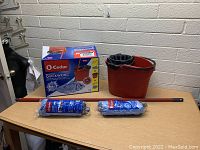 O'Cedar mop and bucket set with mop handle, bucket with wringer, two packaged mop heads, and box displayed on wooden table against white brick wall.