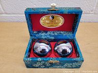 Two shiny silver-tone metallic Baoding balls placed inside an open blue fabric covered case with floral patterns. The interior lining is red fabric with a gold label on the inside lid showing yin-yang symbol and Chinese text.