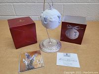 Front view of the white porcelain Lladro Christmas ornament hanging on metal stand with original box and certificate card in background.