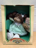 Full view of 1984 Cabbage Patch Kids doll seated inside original box. Shows dress, shoes, box design, and birth certificate visible behind doll.