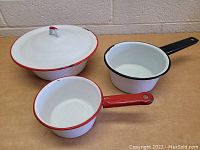 Three vintage enamel pots displayed on wooden surface against a brick wall, showing overall set with handles and cover.