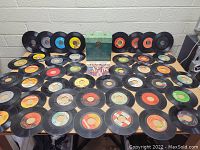 Wide view showing green metal record case surrounded by numerous 45 RPM vinyl records laid out on table