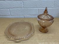 Two vintage pink glass items on a wooden surface against a light grey brick background. Left is a rectangular platter with handles and decorative edges; right is a footed pedestal dish with lid and ornate floral embossing.