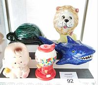 Five ceramic coin banks arranged on a shelf: turtle, lion, baby, gumball machine, shark.