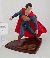 Front and angled views of Kotobukiya Man of Steel Superman statue in blue suit, red boots, and posable red fabric cape on brown S-shaped stand.