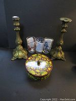 Pair of ornate metal Art Nouveau style candlesticks, one missing wax drip rim, three decorated Hazel Atlas drinking glasses, and vintage floral round Daher tin with lid.