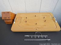 Wide view of wooden carving board with metal handles and pegs along with CUTCO cutting block and metal utensils.