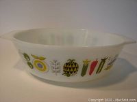 Side view of the white glass round casserole with colorful stylized fruit and vegetable patterns showing green apple, yellow rings, and other motifs.