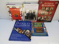 Lot of five Antiques Roadshow related books displayed on a white surface: one with a red cover titled Antiques Roadshow Guide, another with a blue cover celebrating 21 years, a yellow and green covered Behind the Scenes book, the Hidden Treasures Keno Brothers book with two men on the cover, and the red and white Primer book with images of antiques.