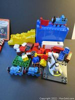 Photo showing multiple LEGO Duplo and Mega Bloks pieces including 3 blue Thomas trains, Harold helicopter with helipad, red flag, yellow ramp, and assorted building blocks.