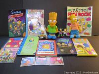 Photo showing complete lot including two Bart Simpson dolls, various books, DVDs, CDs, trading cards, and small toys all arranged neatly on a black background.