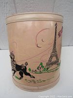 Photo showing the full front side of the cylindrical metal waste basket with pink and gray wash paint, decorated with a black poodle and Eiffel Tower illustration.