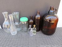Photo showing all 10 glass items: 4 brown glass bottles of varying sizes including 1 large jug, 3 clear glass vases in different shapes, 3 small bottles (2 antique empty, 1 liquid-filled), and 1 vintage Ball mason jar with green lid.