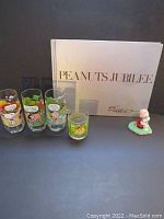 Full lot view showing Peanuts Jubilee book, four drinking glasses, and ceramic Charlie Brown figurine on grass base.
