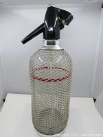 Full view of vintage glass seltzer bottle with silver mesh casing and black dispensing head
