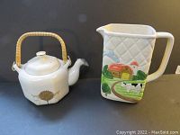 Side view of ivory ceramic teapot with golden tree decoration and rattan handle, plus white ceramic pitcher with cow and barn design.