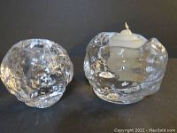Pair of clear glass Kosta Boda snowball-shaped votive candle holders, one containing a white candle, displayed on black surface.