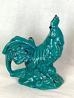 Full view of turquoise ceramic rooster figurine showing side and base with flower details.