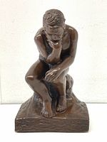 Front view of a seated male figure sculpture in a thinking pose with bronze-like finish, detailed hair and musculature, on a textured square base.