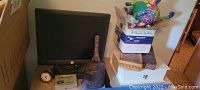 Photo showing a desktop monitor, antique metal cow bell, vintage style clock, boxes and assorted items on the floor and shelf.