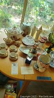 Photo showing an assortment of pottery, glass, crystal, vases, teacups and figurines arranged on a wooden table near window light.