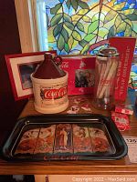 Photo showing Coca Cola metal serving tray with vintage artwork, ceramic jug cookie jar with Coca Cola label, clear Coca Cola straw dispenser with straws, red Coca Cola tray, and framed polar bear print.