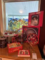 Wide view of assorted Coca Cola collectibles on wooden shelf with stained glass window background. Items visible include tins, pitcher, glasses, playing cards, and boxed platter.