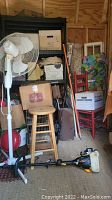 Photo of lot showing white stand fan, wooden stool with padded seat, red ladder back chair, gas-powered string trimmer, and storage boxes on shelves.