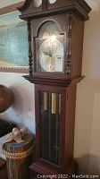 Front view of grandfather clock showing wooden case, clock face with Roman numerals, and pendulum compartment with weights behind glass door.