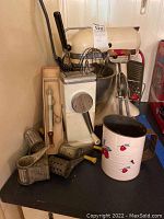 Photo showing vintage KitchenAid cream-colored stand mixer with bowl, a hand rotary mixer with attachments, thermometer, and grater utensils on a black surface.