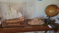 Two handcrafted matchstick ships and a vintage globe on wood surface. One ship inside a clear display case.