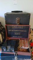 Black textured carrying case on top. Below it, a wooden box with 'Zealand Royal Casket Company' branding opened, containing the realist viewfinder and slide collection.