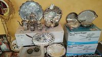Lot of silver-plated serving ware including coffee pots, trays, creamer, sugar bowls, and serving platters, arranged on and around boxes.