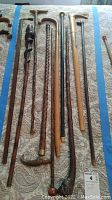 Ten vintage wooden canes laid out on fabric, showing various handle designs including mother of pearl and silver accented handles, carved and textured wood shafts.