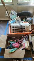 Photo showing box fan heater, colorful decorative bird wall hanging, and miscellaneous small household items in boxes including a pink decorative mask.
