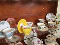 Overview of the 10 vintage teacups and saucers with floral designs arranged on glass shelf.