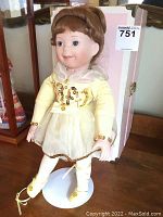 Porcelain ballerina doll dressed in yellow and gold ballet outfit standing on white circular stand inside glass display case. Doll has brown braided hair and blue eyes.